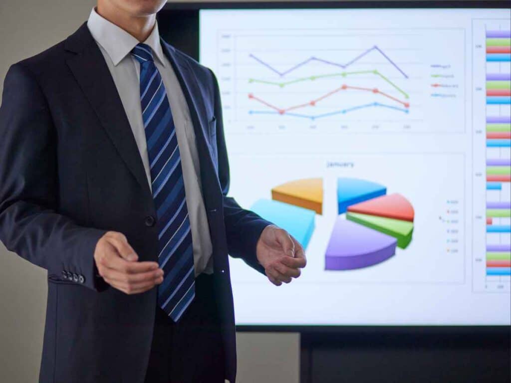Homem de terno fazendo uma apresentação profissional com gráficos de pizza e linhas exibidos em uma tela, representando o uso do PowerPoint