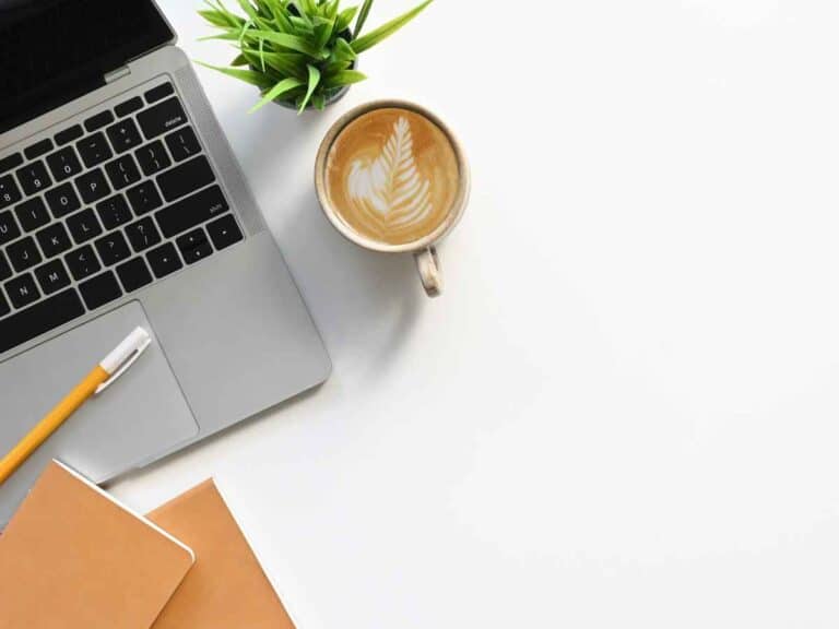 Mesa de trabalho com notebook aberto, caderno, caneta, xícara de café com arte no leite e vaso pequeno de planta