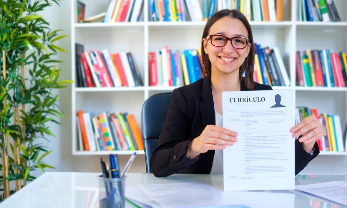 mulher exibindo um currúculo, sentada em sua mesa com uma estante de livros no fundo a imagem