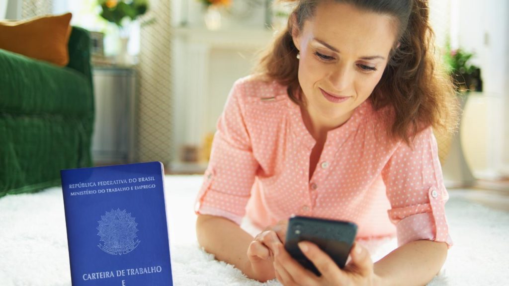 mulher mexendo no celular para consulta do valor do seguro-desemprego, e uma carteira de trabalho ao lado, na imagem