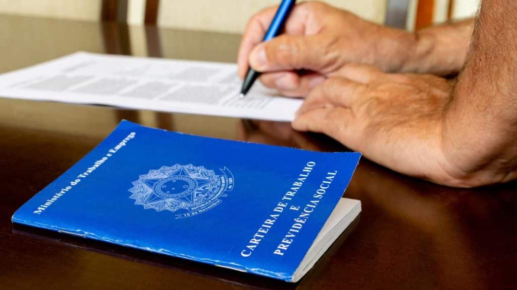 carteira de trabalho e ao lado uma pessoa assinando um documento, sobre seguro-desemprego, em cima de uma mesa