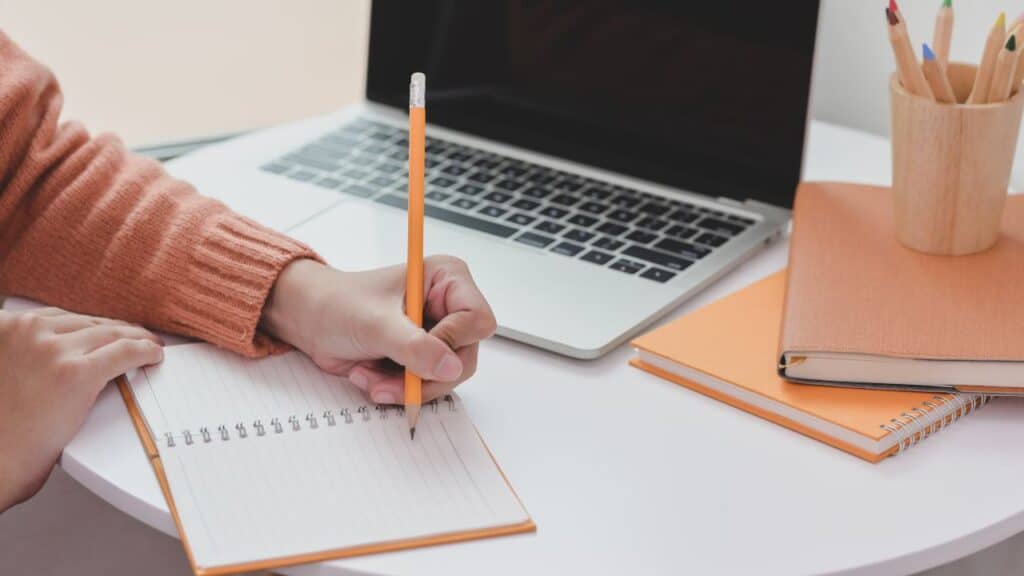 mulher escrevendo em um caderno, em sua mesa está o computador e os cadernos.