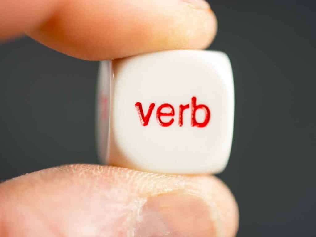 Dado branco com a palavra “verb” escrita em vermelho, sendo segurado por uma pessoa
