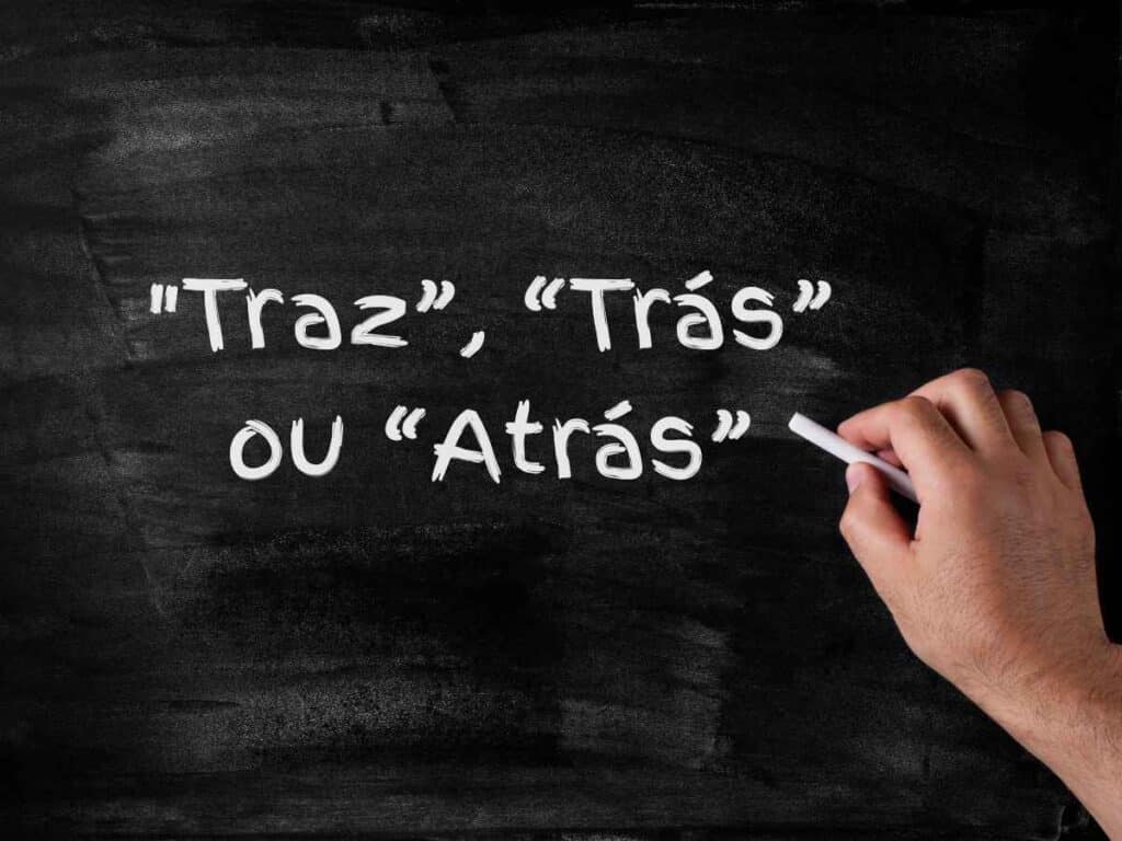 Mão escrevendo em uma lousa preta com giz branco as palavras “Traz”, “Trás” ou “Atrás”