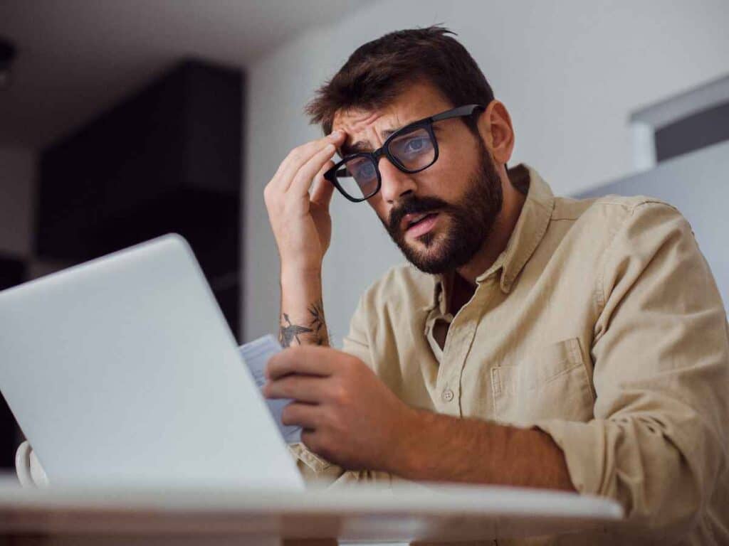 Ex-beneficiário do seguro-desemprego vendo que seu benefício foi cancelado e que terá que devolver o valor das parcelas recebidas. Ele observa o computador com um olhar incrédulo, enquanto apoia sua cabeça.