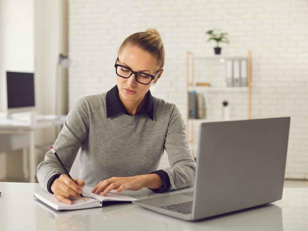 Beneficiária do seguro-desemprego sentada à mesa, participando de um curso online com o objetivo de melhorar suas chances no mercado de trabalho, adquirir novos conhecimentos e se desenvolver pessoal e profissionalmente. À sua frente, um notebook exibe o conteúdo do curso enquanto ela faz anotações em um caderno com foco e dedicação.