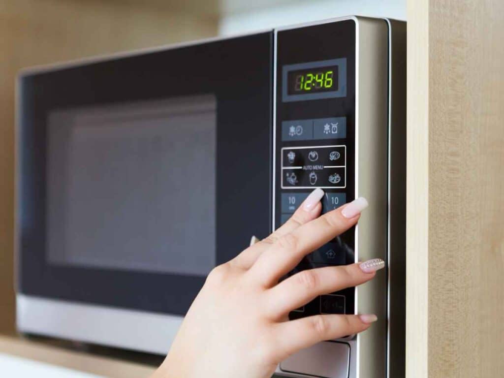 Mão de uma mulher acionando o painel de controle de um micro-ondas, ligando o aparelho durante o uso. Enquanto ela se questiona mentalmente sobre a forma correta de escrever o nome desse utensílio.