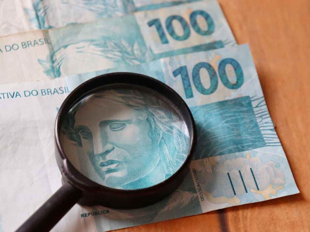 Lupa ampliando o rosto da efígie da República em uma cédula de 100 reais, com outras notas do mesmo valor ao fundo, sugerindo análise minuciosa de autenticidade ou avaliação de cédulas.