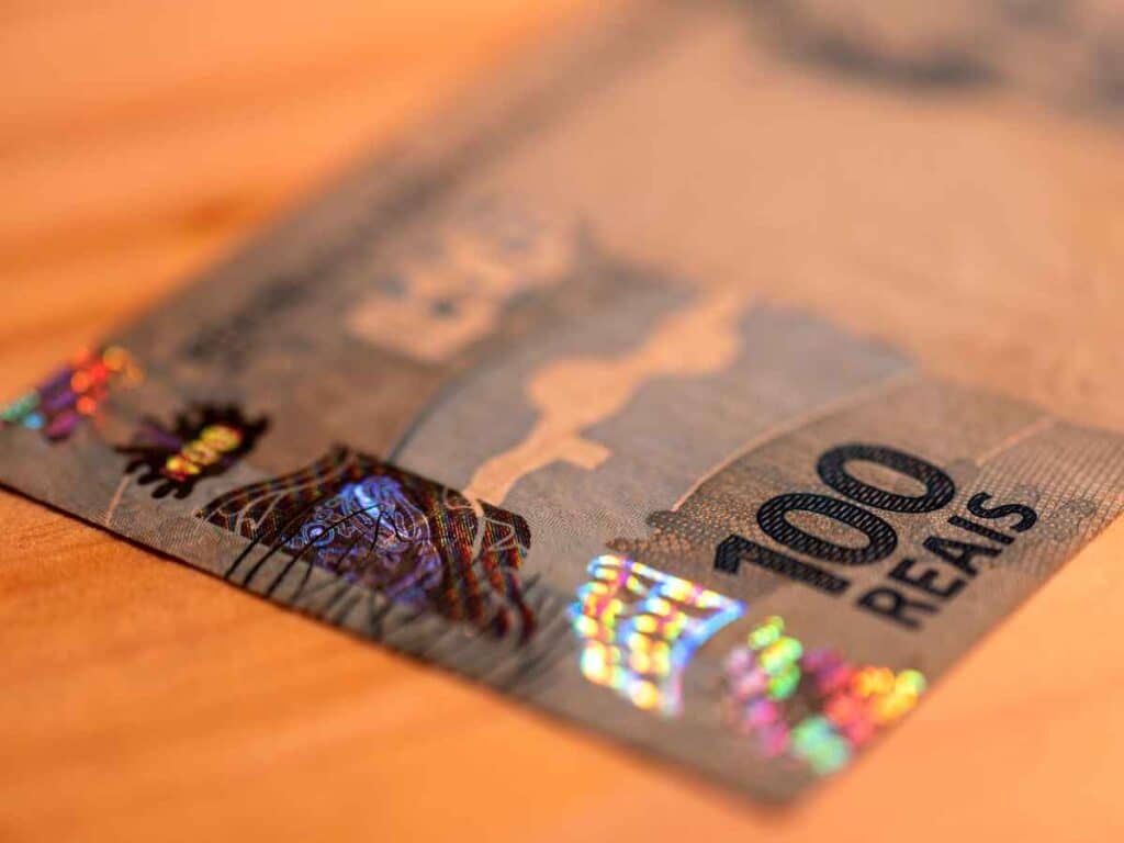 Close-up de uma cédula de 100 reais com foco na faixa holográfica de segurança, destacando os elementos brilhosos e coloridos usados para prevenir falsificações.