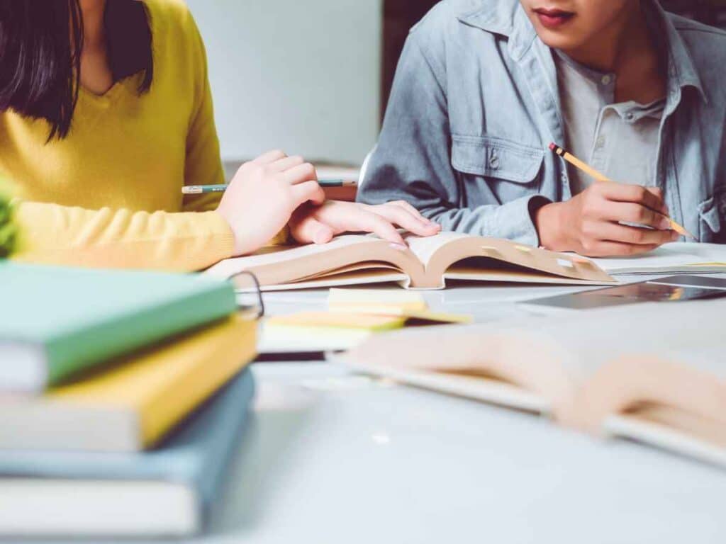 Dois estudantes sentados lado a lado em uma mesa, concentrados na realização de exercícios de português. Eles escrevem com lápis e consultam livros abertos, rodeados por cadernos, livros empilhados e post-its.