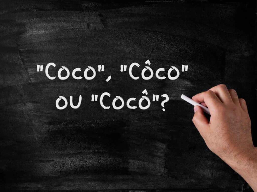 Mão segurando giz branco em frente a uma lousa preta com a frase escrita: "Coco", "Côco" ou "Cocô"?