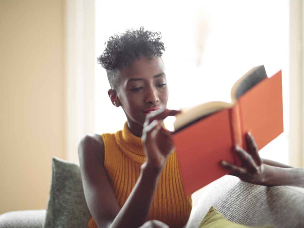Mulher sentada em um sofá, concentrada na leitura de um livro de capa laranja. Ela usa uma blusa sem mangas de gola alta na cor mostarda e está em um ambiente iluminado pela luz natural que entra pela janela ao fundo.