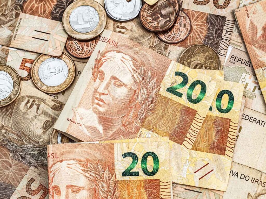 Notal e moedas do real espalhadas, com destaque para a cédula de 20 reais posicionada em evidência no centro da imagem.
