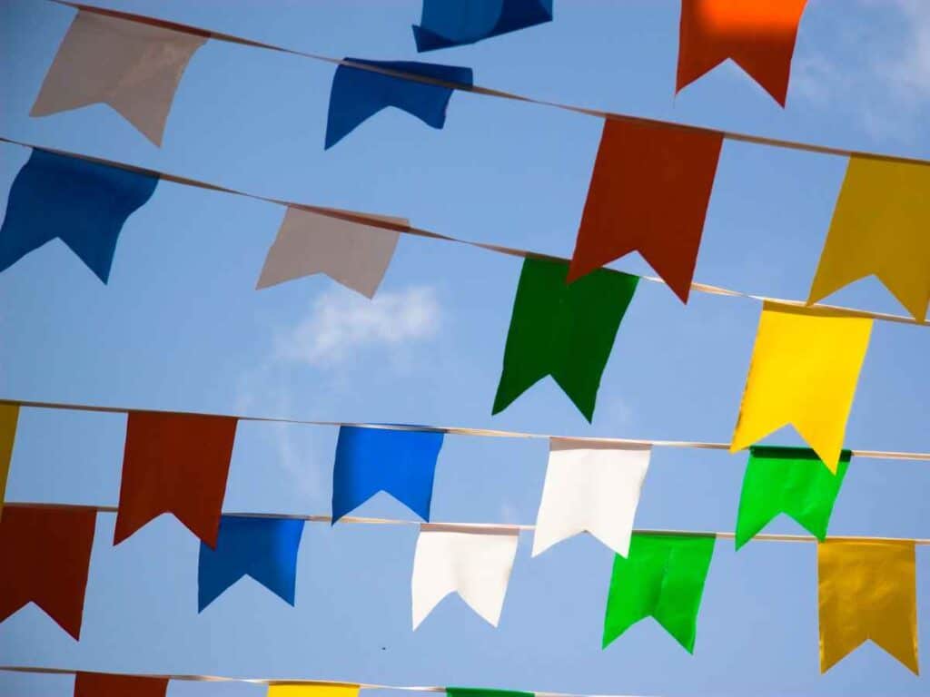 Fileiras de bandeirinhas coloridas típicas de festa junina, penduradas sob um céu azul claro com algumas nuvens.