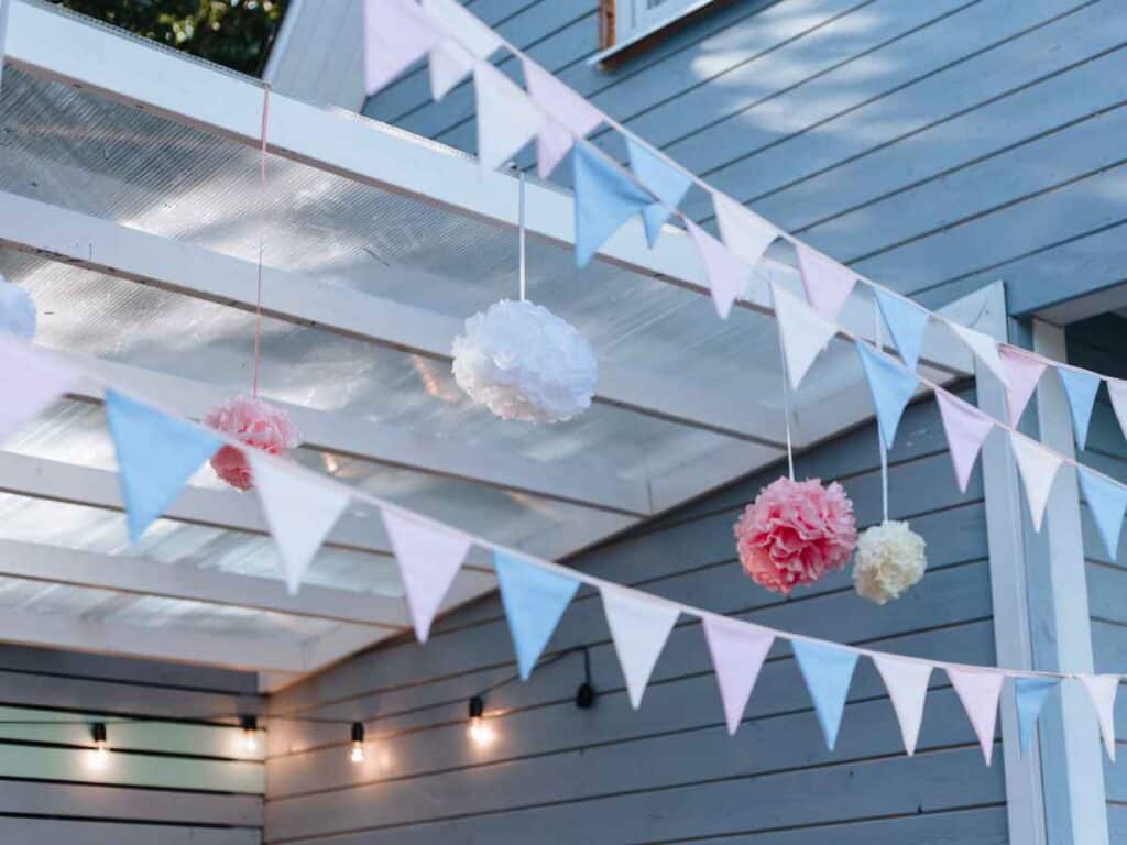 Bandeirolas triangulares em tons pastéis de azul, rosa e branco decoram uma varanda com estrutura branca e parede azul, junto a pompons de papel pendurados, criando um clima delicado e festivo, ideal para celebrações de Réveillon.