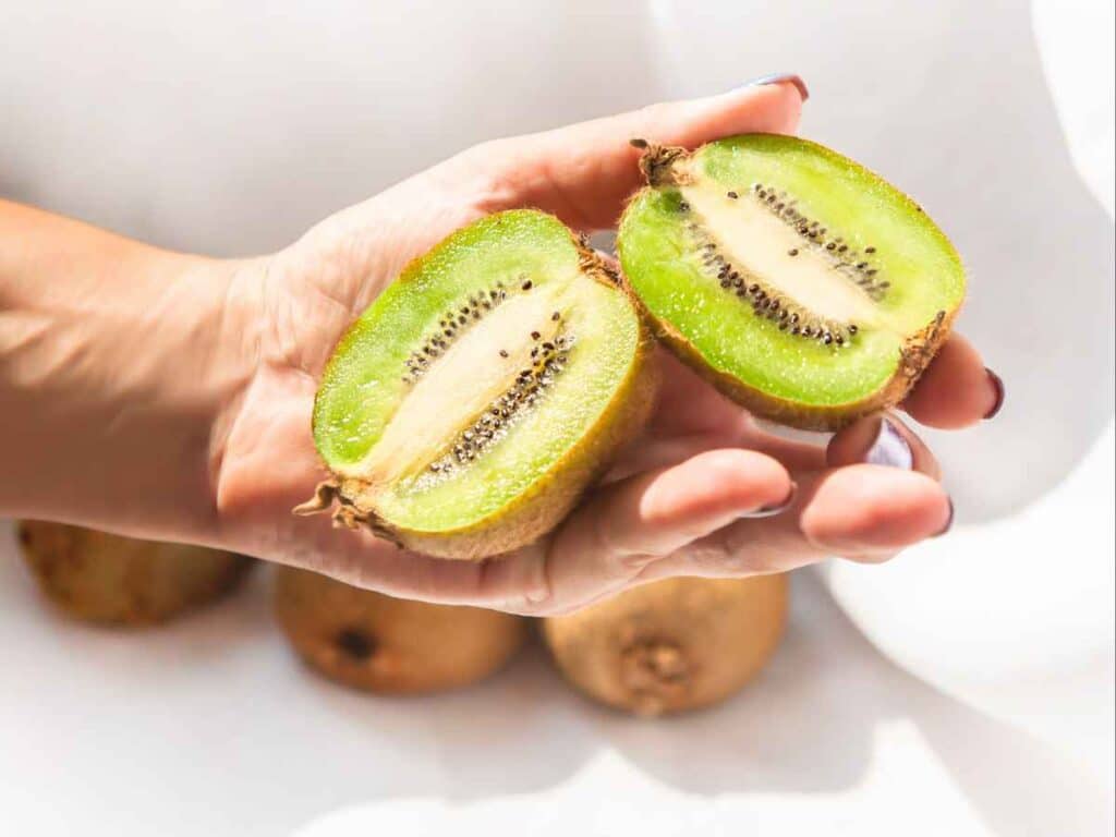 Mão segurando um kiwi partido ao meio, exibindo sua polpa verde vibrante e sementes escuras.