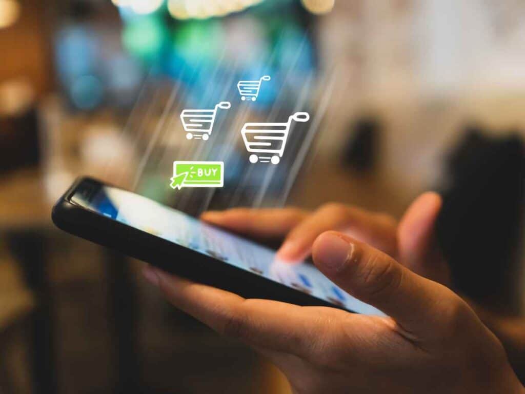 Pessoa segurando um celular com ícones digitais de carrinhos de compra e botão “buy”, representando o conceito de transações online, compra e venda de moedas