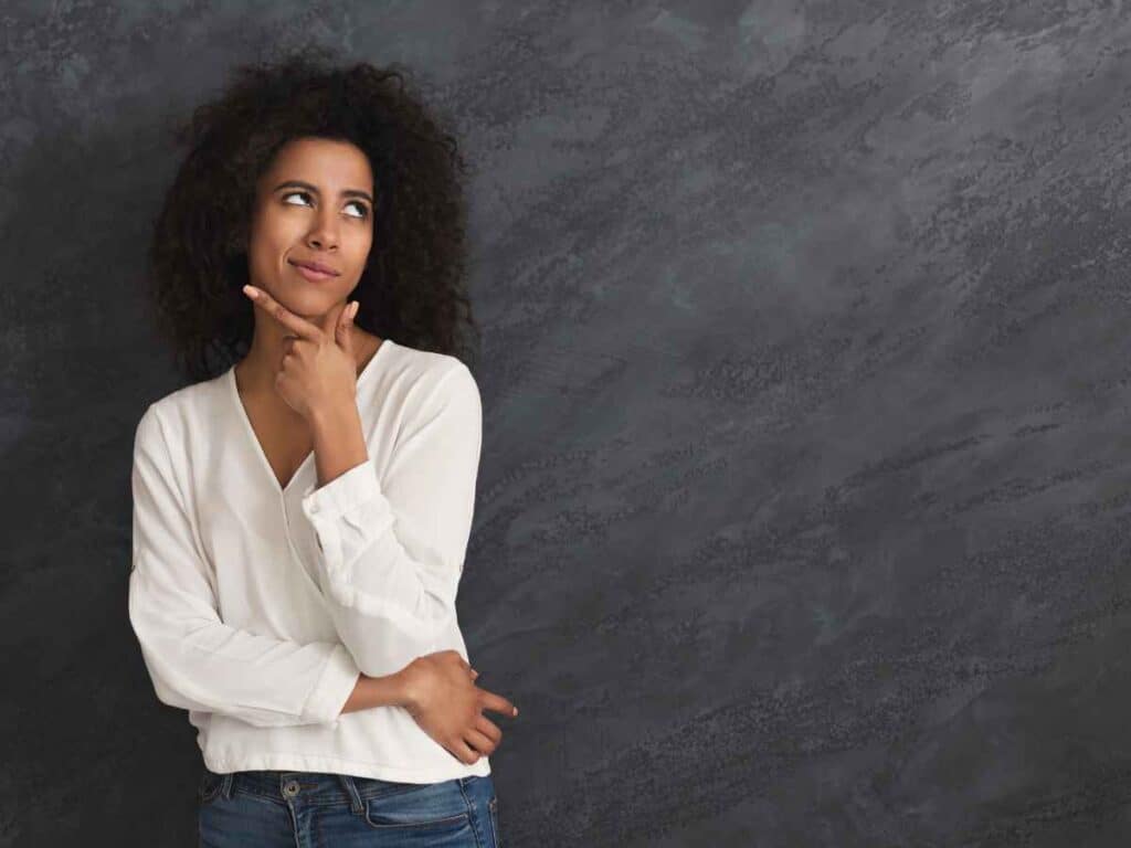 Mulher jovem com expressão de dúvida, levando a mão ao queixo enquanto olha para cima, em frente a um fundo cinza escuro