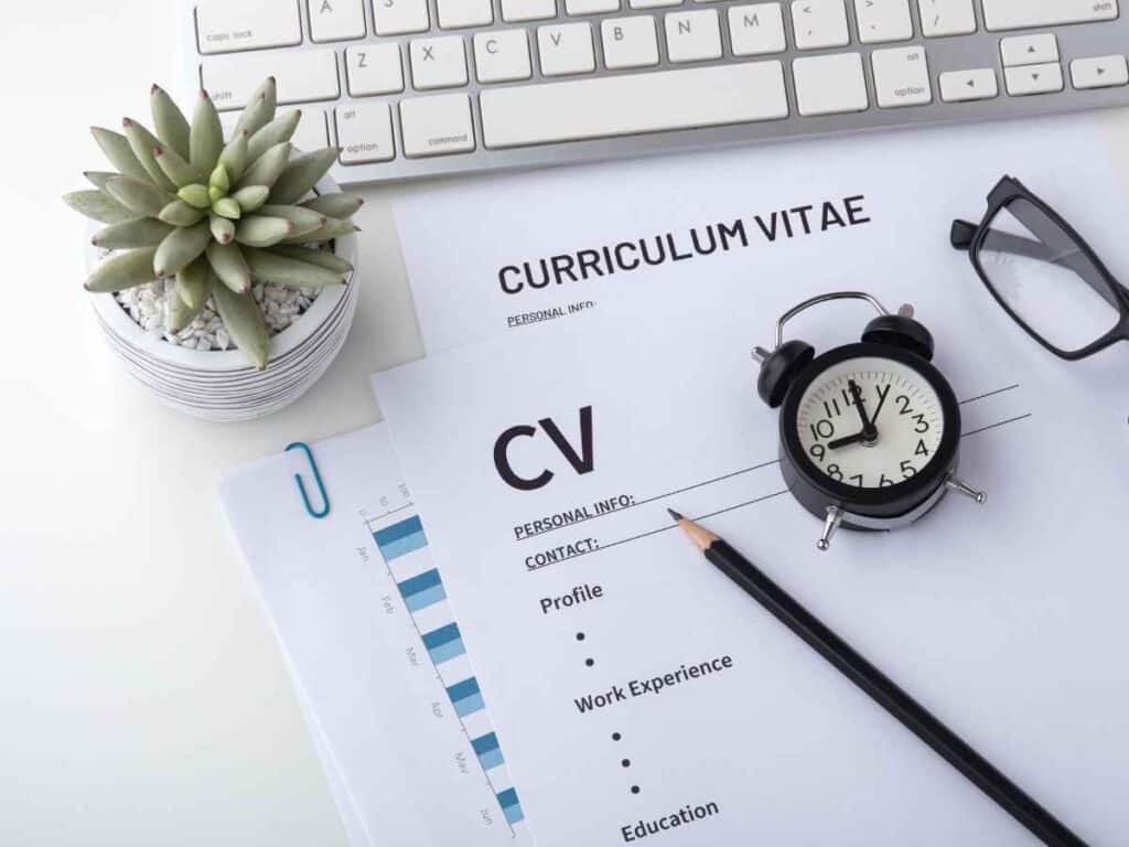 Currículo sobre mesa com relógio, lápis, óculos e teclado, destacando organização e planejamento para buscar emprego.