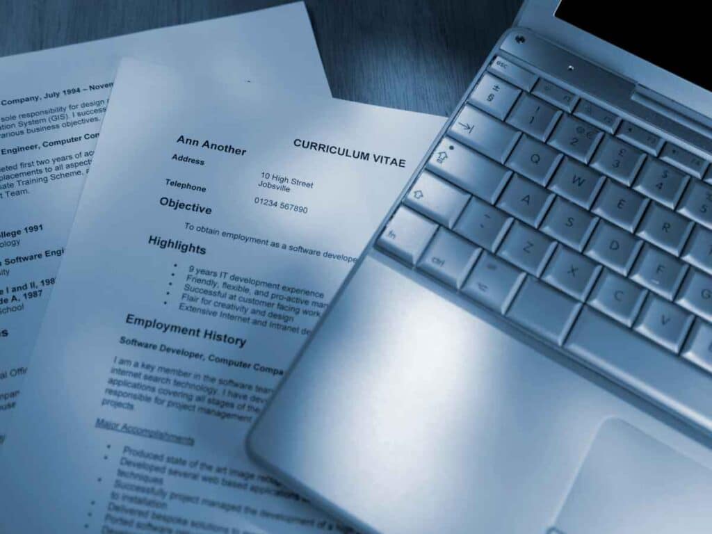 Currículo impresso ao lado de um notebook, representando preparação para candidatura a vaga de emprego.