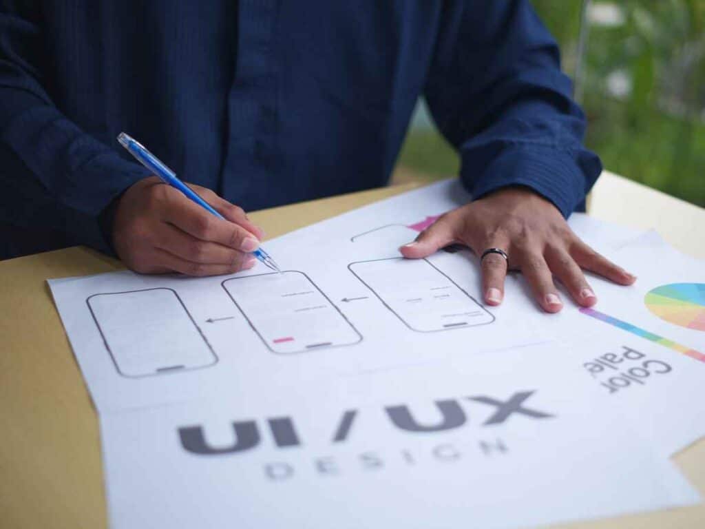 Pessoa desenhando wireframes de aplicativo em uma folha com o texto UI/UX Design, representando planejamento de interface.