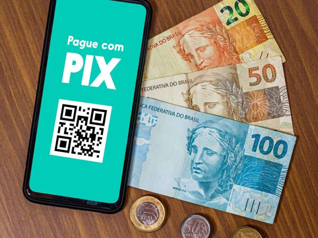 Tela de celular exibindo “Pague com PIX” ao lado de cédulas de real e moedas sobre uma mesa.