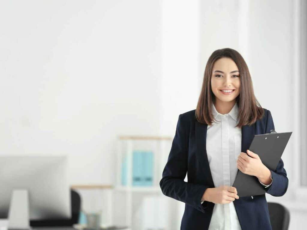 Mulher sorrindo em ambiente corporativo segurando uma prancheta, representando organização e perfil profissional.