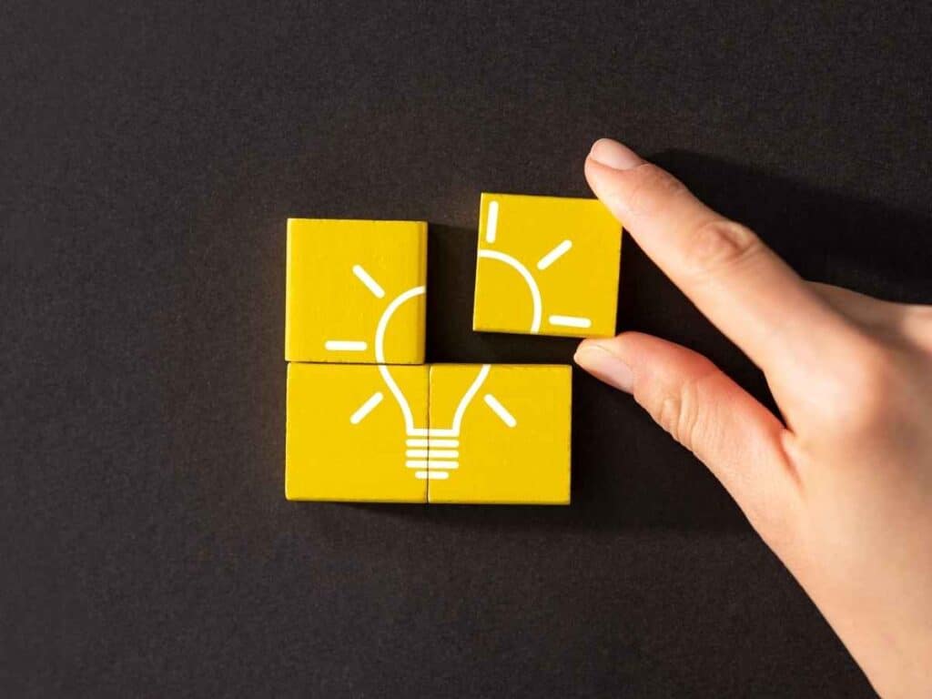 Blocos amarelos formando o desenho de uma lâmpada sobre fundo preto, com uma mão posicionando uma das peças na parte superior.