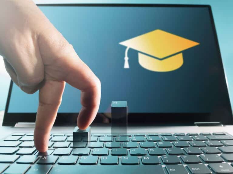 Mão pressionando teclas de um notebook, com ícone de chapéu de formatura na tela, representando ensino e aprendizado online com certificado.