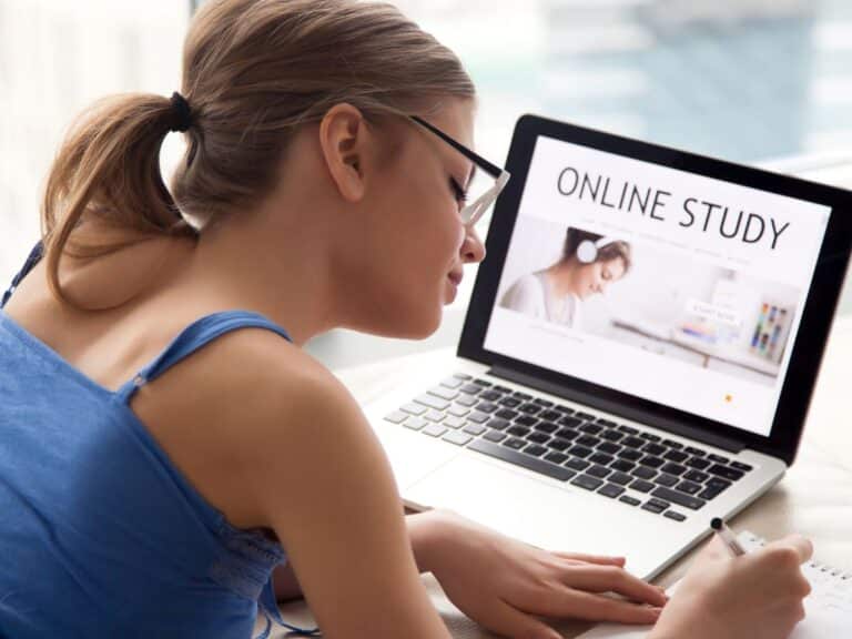 Mulher estudando em casa com notebook aberto em uma plataforma de estudo online, fazendo anotações em um caderno ao lado do computador.