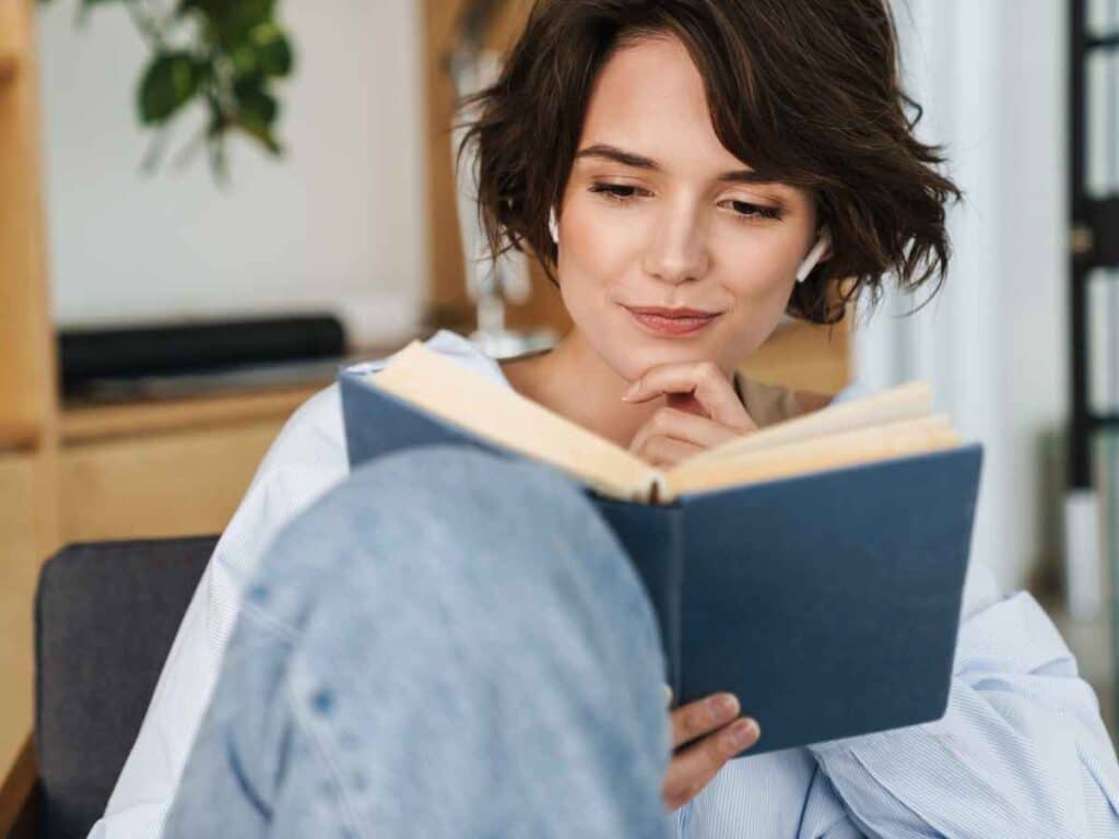Mulher sentada lendo um livro de capa azul em ambiente interno, com expressão concentrada.