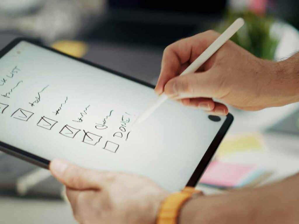 Mãos segurando um tablet enquanto uma caneta digital marca itens de uma lista manuscrita na tela.