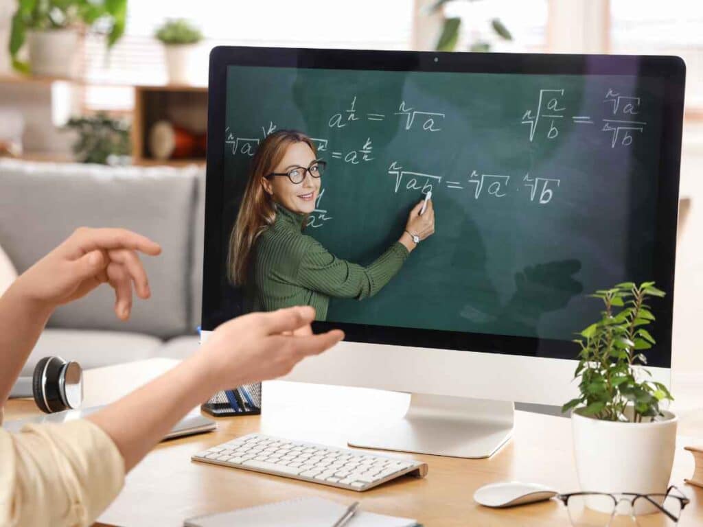 Monitor de computador exibindo professora ensinando matemática em quadro, enquanto uma pessoa acompanha a aula online e gesticula durante o aprendizado em casa.