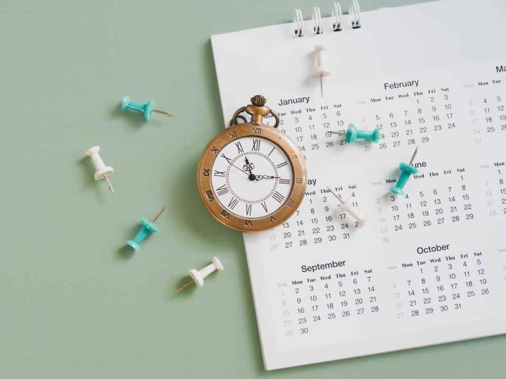Calendário de mesa com meses visíveis ao lado de um relógio analógico e alfinetes coloridos, representando planejamento