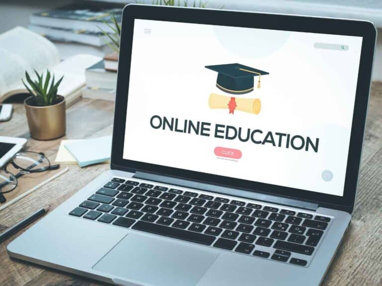 Notebook sobre mesa de madeira exibindo a frase “online education” na tela, cercado por materiais de estudo como caderno, óculos e planta decorativa.