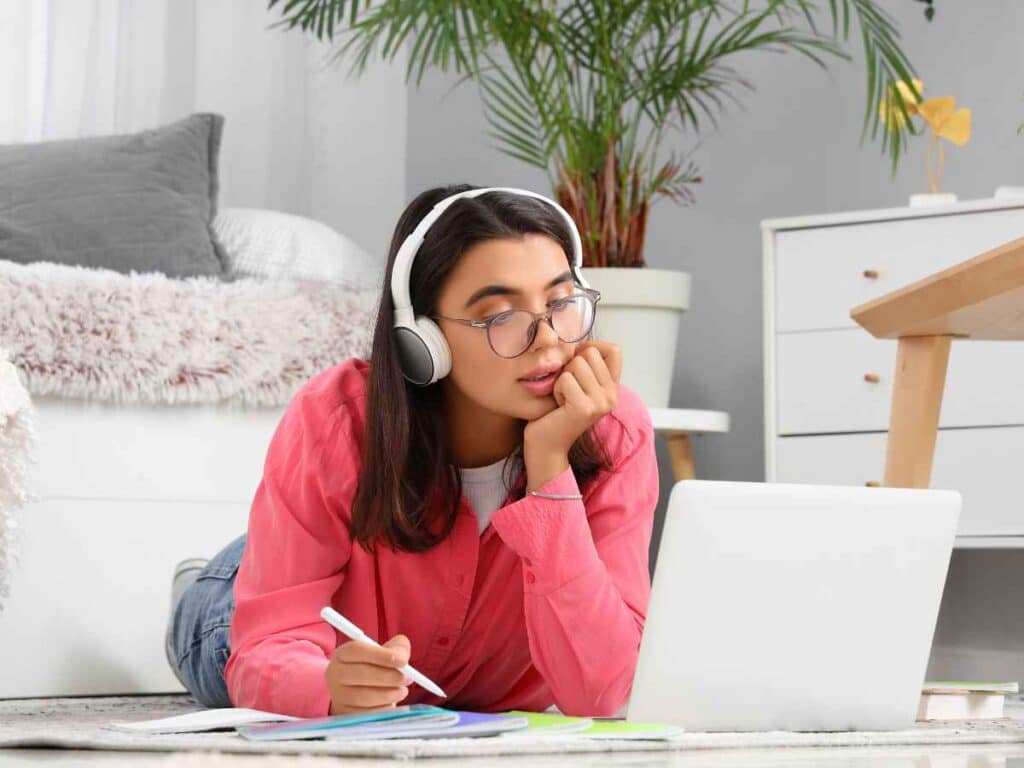 Jovem deitada no chão com fones de ouvido utilizando notebook e escrevendo em caderno, representando estudo online em ambiente confortável.