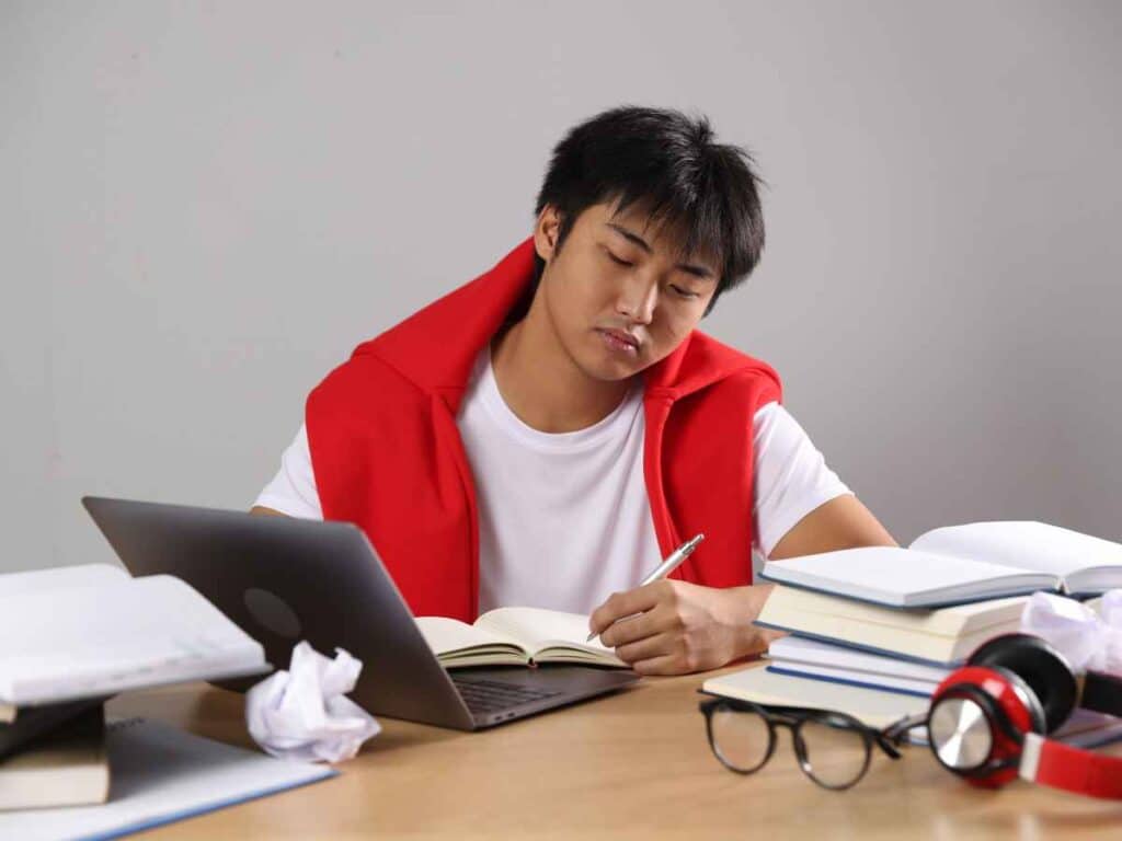 Jovem estudando em mesa com notebook e livros empilhados, escrevendo em caderno enquanto se dedica à organização dos estudos e tarefas profissionais