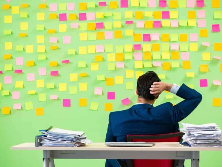 Homem sentado de costas em frente a uma parede coberta por diversos post-its coloridos, aparentando analisar e organizar ideias e tarefas
