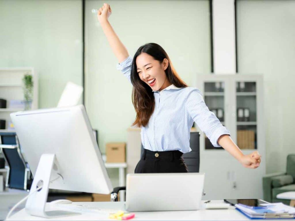 Mulher em escritório levantando os braços com expressão de alegria em frente ao computador, celebrando conquista