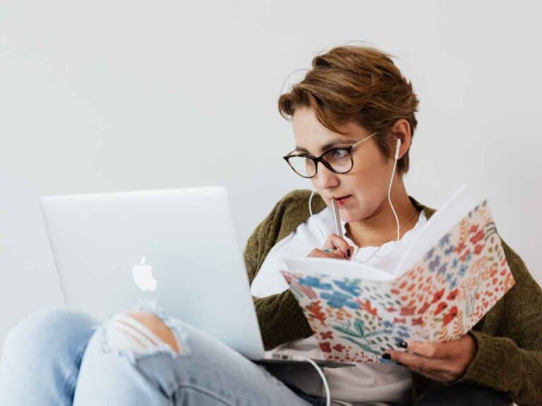 Mulher concentrada estudando online, o notebook esta apoiado em seus joelhos e ela faz anotações em um caderno florido
