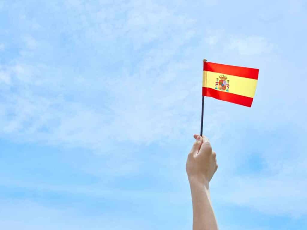 Mão segurando uma pequena bandeira da Espanha diante de um céu azul com poucas nuvens
