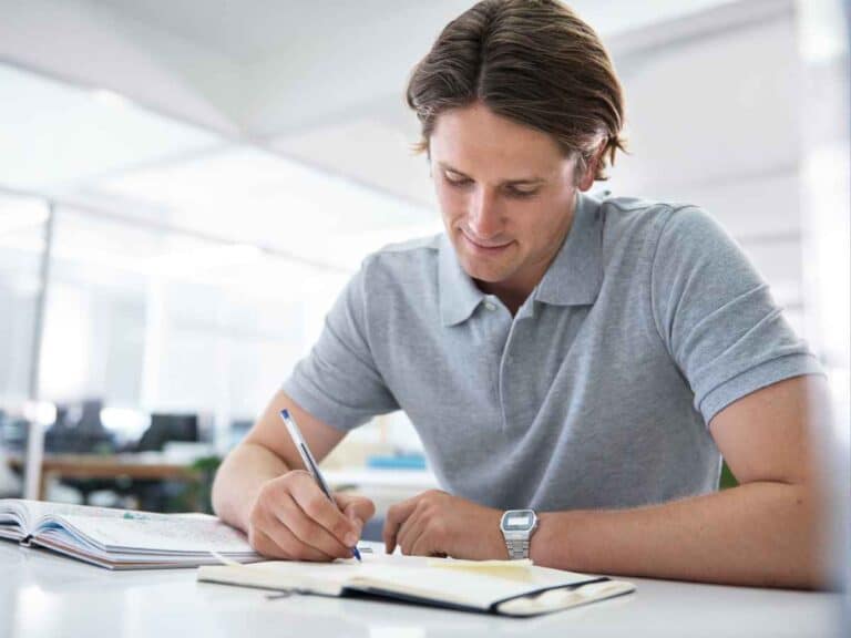 Homem sentado à mesa escrevendo em um caderno durante os estudos, com livros e materiais de apoio ao lado, em ambiente claro e organizado.