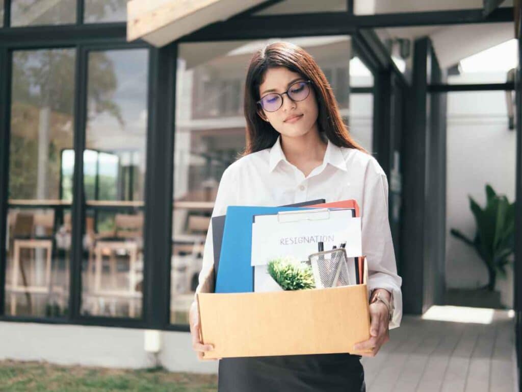 Mulher sai de um ambiente corporativo carregando uma caixa com documentos e objetos pessoais