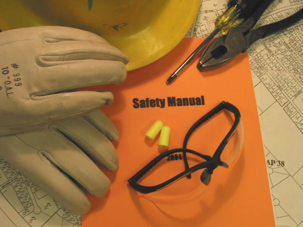 Capacete de segurança, luvas de proteção, óculos de segurança, protetores auriculares e ferramentas dispostos sobre um manual de segurança do trabalho