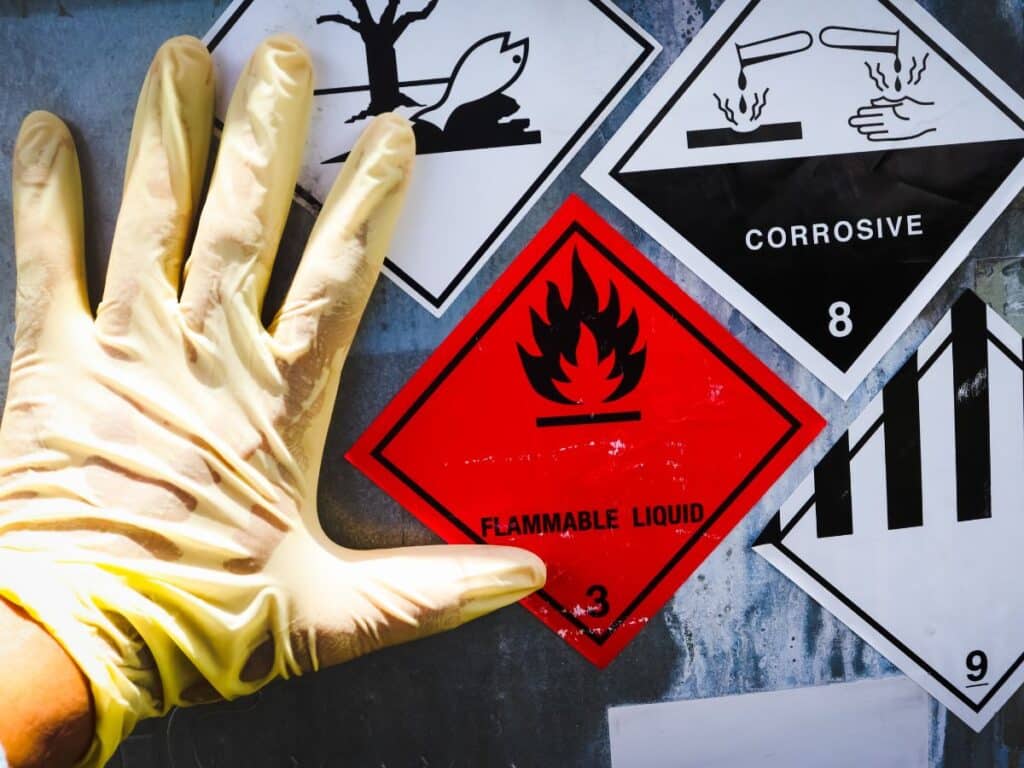 Luva de proteção posicionada sobre placas de sinalização de riscos químicos e materiais inflamáveis