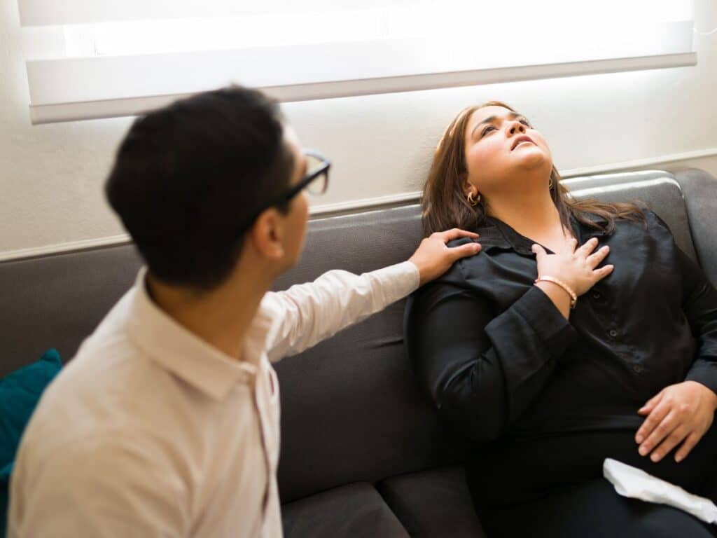 Pessoa oferece apoio a mulher durante uma crise de ansiedade, tocando seu ombro enquanto ela respira profundamente sentada no sofá.