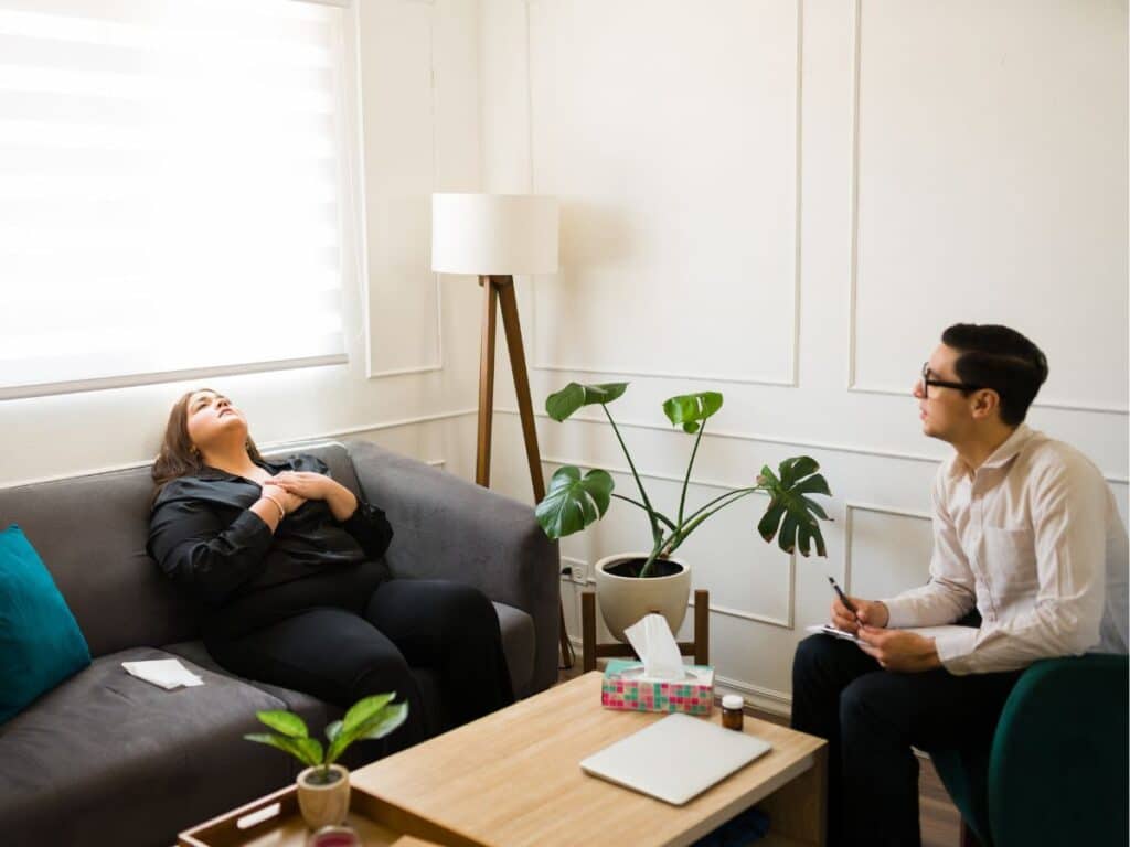 Mulher conversa com psicólogo durante atendimento, sentada no sofá com as mãos sobre o peito enquanto recebe orientação profissional.